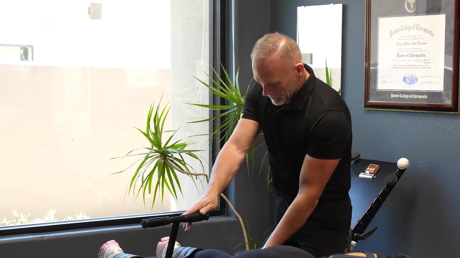 Dr. Steve Van Laecken DC at a treatment table with a patient lying face down, his Parker College of Chiropractic diploma on the wall behind him